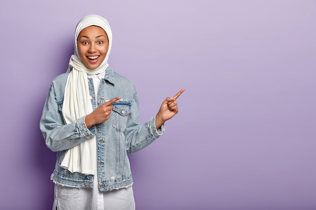 Photo gratuite heureuse femme métisse a une expression joyeuse, pointe du doigt avec les deux doigts avant, dit suivre là, montre la direction sur l'espace de copie, porte des vêtements en jean, hijab blanc, isolé sur un mur violet