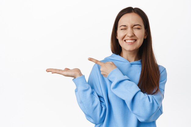 Heureuse femme mignonne en sweat à capuche, riant et souriant, pointant le doigt vers la paume ouverte vide, montrant quelque chose dans sa main, debout contre un mur blanc