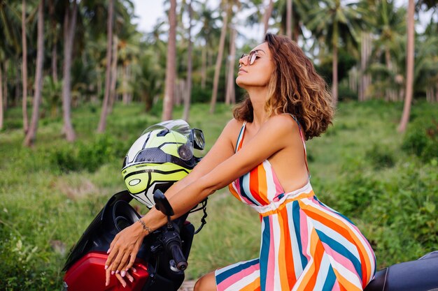 Heureuse femme de motard caucasien en robe d'été colorée en vacances avec casque de moto