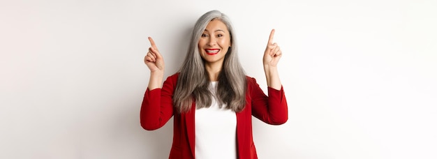 Photo gratuite heureuse femme mûre en blazer rouge et maquillage souriant et montrant la publicité sur le doigt pointé du haut