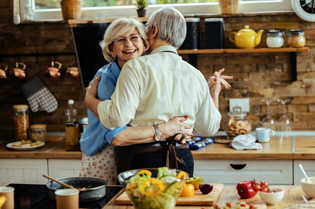 Heureuse femme mûre et son mari dansant tout en préparant la nourriture dans la cuisine