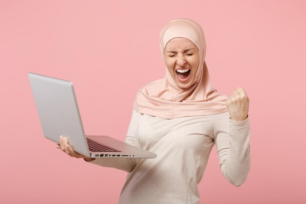 Heureuse femme musulmane arabe en vêtements légers hijab posant isolé sur fond rose. concept de style de vie religieux des gens. maquette de l'espace de copie. tenez l'ordinateur portable, faites le geste du gagnant, hurlez.