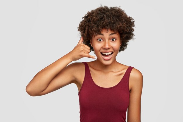 Heureuse femme noire avec une coiffure afro fait un geste de téléphone avec la main comme parler sur cellulaire