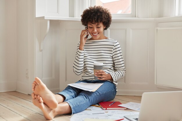 Heureuse femme noire avec une coupe de cheveux afro, parle par téléphone portable tout en examinant des documents