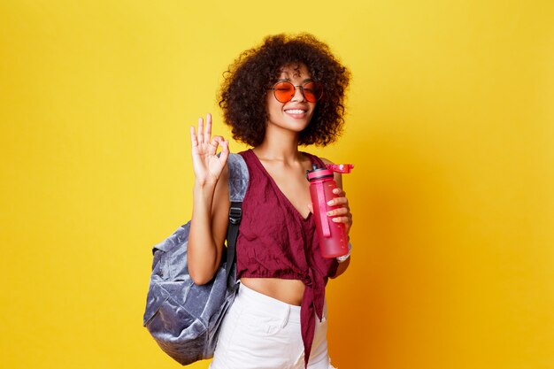 Heureuse femme noire ludique en tenue d'été élégante avec signe de paix qui pose en studio sur fond jaune. Tenant une bouteille d'eau. Coiffure afro. Mode de vie sain.