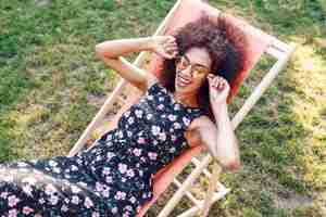 Photo gratuite heureuse femme noire à la mode avec une coiffure frisée élégante assis sur une chaise longue sur une pelouse verte incroyable