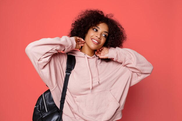 Heureuse femme noire sexy en sweat à capuche élégant avec sac à dos posant sur fond rose et jouant avec des cheveux bouclés.