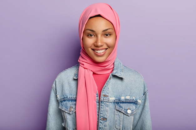 Heureuse femme orientale a la religion islamique, la tête couverte de voile rose, sourit doucement, montre des dents blanches, isolée contre le mur violet exprime des sentiments et des émotions positifs. Ethnicité