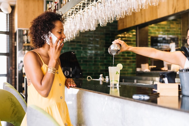 Heureuse femme parlant sur téléphone mobile en regardant barman faisant un cocktail au bar