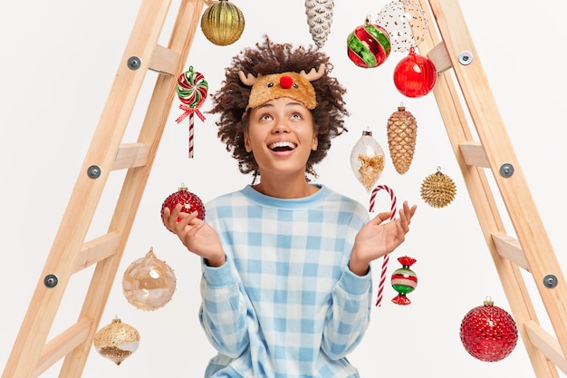 Heureuse femme à la peau foncée regarde positivement ci-dessus sur les boules et les jouets du Nouvel An vêtus d'un pyjama et d'un masque de sommeil se propage les paumes apprécie les vacances d'hiver décore la maison bénéficie d'une atmosphère domestique