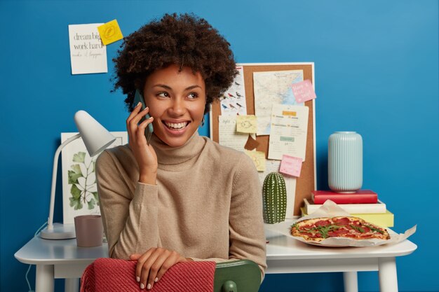 Heureuse femme à la peau sombre a une conversation téléphonique, regarde ailleurs, étant de bonne humeur comme travail terminé, s'assoit contre le bureau avec une pile de cahiers, des notes autocollantes sur le mur et le tableau, une délicieuse pizza