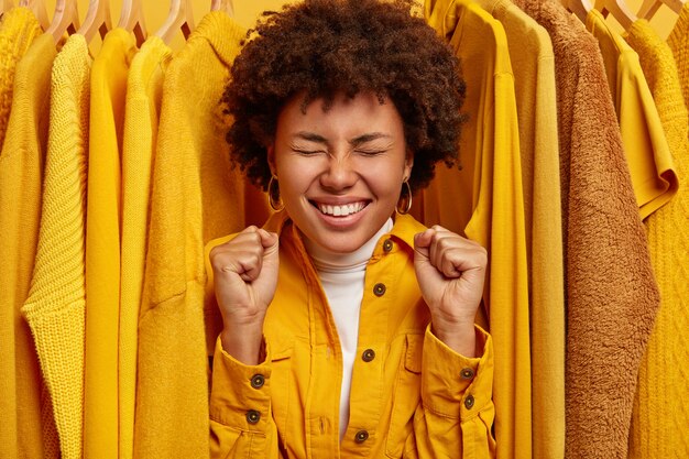 Heureuse femme à la peau sombre ravie se tient près de vêtements élégants jaunes sur des cintres, serre les poings, se réjouit de l'achat réussi