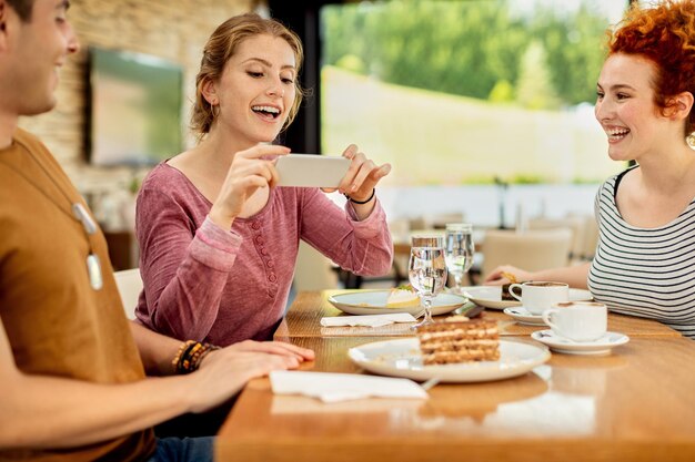 Heureuse femme photographiant un dessert avec un téléphone portable tout en étant avec ses amis dans un café