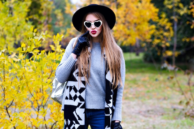 Heureuse femme porte un manteau élégant et un chapeau noir posant dans des lunettes de soleil en journée d'automne ensoleillée. Portrait en plein air de modèle féminin enthousiaste en pull gris à la mode marchant dans le parc avec des arbres jaunes.