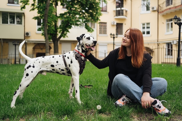 Heureuse femme posant et jouant avec son chien dalmatien assis dans l'herbe verte lors d'une promenade en ville