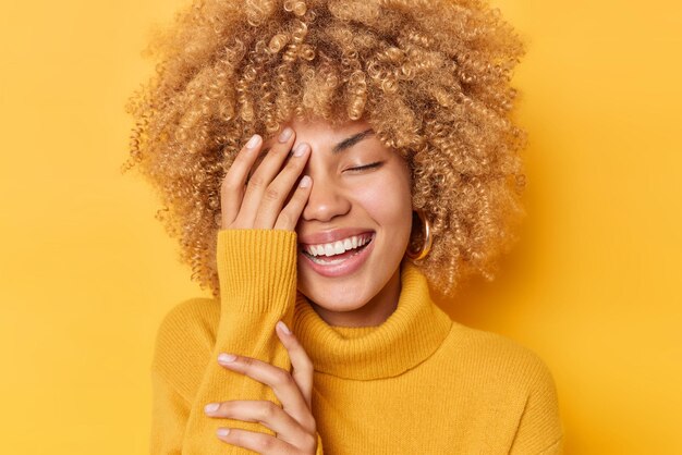 Heureuse femme positive avec des cheveux bouclés et touffus fait des sourires de paume de visage montre largement les dents blanches garde les yeux fermés porte un pull confortable isolé sur fond jaune vif Sentiments sincères