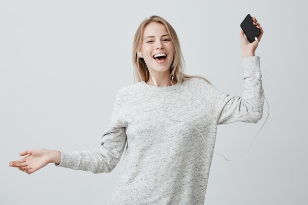 Heureuse femme positive danse dans la joie, tient le téléphone portable, sourit largement, écoute les chansons préférées dans les écouteurs blancs, utilise la musique ap