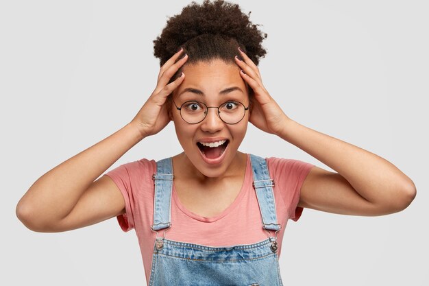 Heureuse femme postive à la peau foncée, garde les mains sur la tête, ne peut pas croire qu'elle a finalement terminé tout le travail, habillée avec désinvolture, pose sur un mur blanc. Surpris femme afro-américaine se demande quelque chose