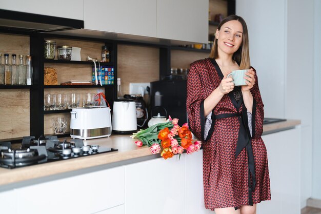 Heureuse femme prenant un café à la maison dans la cuisine portant une robe de soie tout en profitant de fleurs fraîches