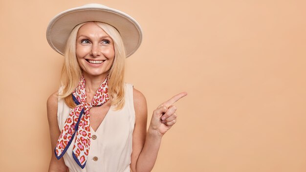 Heureuse femme de quarante ans en robe chapeau élégante et foulard sur le cou indique loin sur l'espace de copie annonce quelque chose montre le chemin du centre commercial isolé sur un mur marron