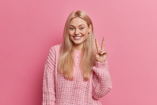 Heureuse femme à la recherche agréable aux cheveux clairs fait signe de paix, sourit à pleines dents