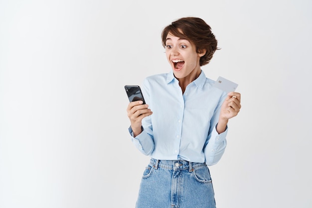 Heureuse femme regardant l'écran du smartphone et criant étonné tenant une carte de crédit en plastique faisant de l'argent en ligne fond blanc