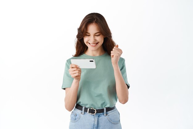 Heureuse femme regardant sur son téléphone portable et se réjouissant de gagner en ligne célébrant la victoire debout avec un smartphone sur fond blanc