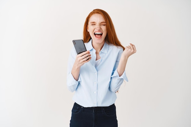 Heureuse femme rousse crie de joie et de satisfaction, remportant un prix en ligne, tenant un smartphone et serrant une main en triomphe, mur blanc