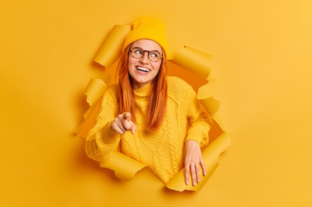 Photo gratuite heureuse femme rousse souriante pointe vers, porte un chandail de chapeau et des lunettes.