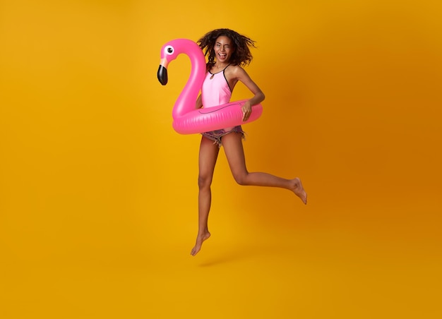 Heureuse femme sautant avec un anneau en caoutchouc flamingo sur fond de fond jaune femme relaxante sur la plage