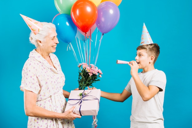 Heureuse femme senior avec cadeau d'anniversaire et fleurs regardant son petit-fils soufflant dans une corne de fête