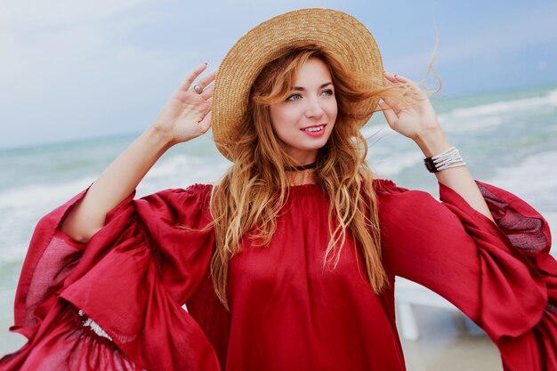 Heureuse femme souriante au gingembre blanc en robe rouge élégante faisant un téléphone mobile autoportrait sur la plage près de l'océan. Porter un chapeau de paille.