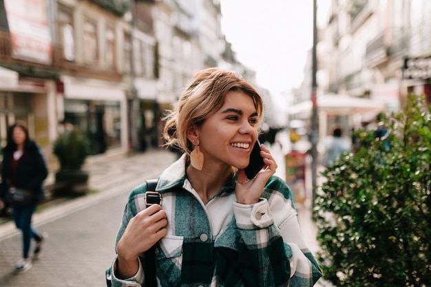 Heureuse femme souriante aux cheveux blonds recueillis, parler sur smartphone dans la rue