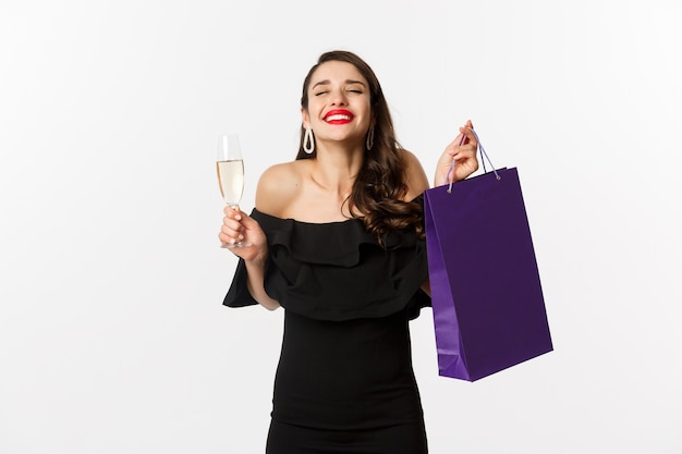 Heureuse femme souriante célébrant, tenant présent dans un sac à provisions et un verre de champagne, debout en robe noire sur fond blanc.