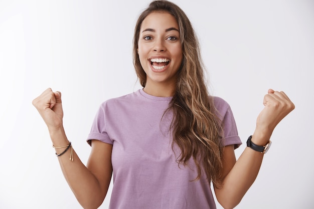 Heureuse femme souriante charmante excitée, cheveux bouclés bronzés, poings serrés célébrant joyeusement l'accomplissement, victoire réussie, hourra, victoire joyeuse, mur blanc amusé debout