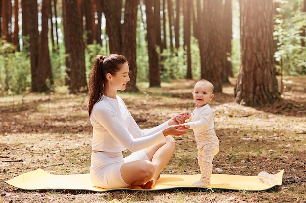 Heureuse femme souriante dans des vêtements de sport à la mode blancs assis sur un tapis de gym en plein air, tenant des palmiers pour enfants