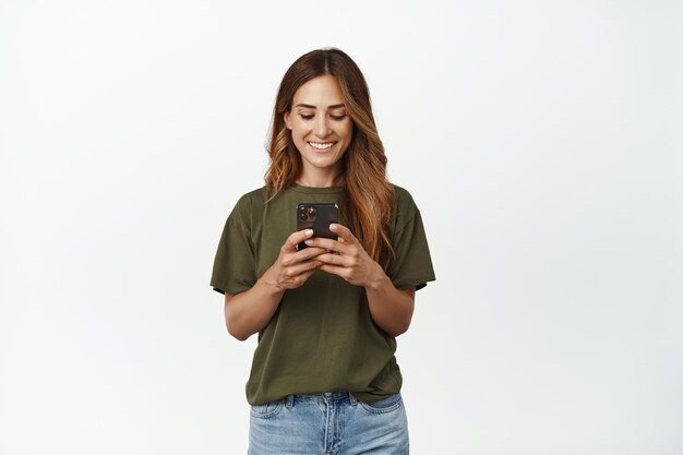 Heureuse femme souriante, femme adulte regardant son téléphone portable, chatter dans l'application, envoyer un message, utiliser l'application d'achat en ligne, debout sur fond blanc.