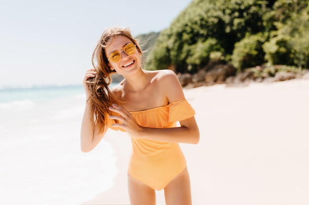 Heureuse femme sportive touchant ses cheveux bruns tout en posant sur la plage de sable. Enchanteur fille caucasienne en maillot de bain jaune s'amuser à l'île tropicale.