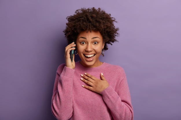 Heureuse femme surprise se sent touchée de recevoir les félicitations d'un vieil ami, bénéficie d'appels gratuits en itinérance, rit pendant les conversations sur smartphone, garde la main sur la poitrine