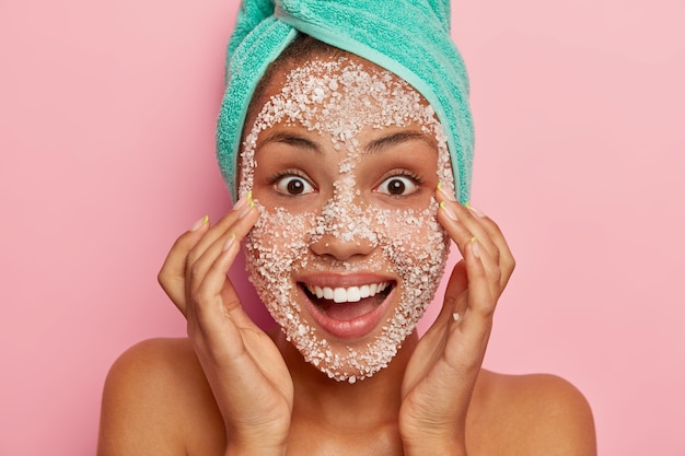 Heureuse Femme Surprise Veut Rafraîchir La Peau Du Visage, Pèle Avec Un Masque De Sel De Mer Naturel, Sourit Paysanement Sourit à La Caméra A Largement Ouvert Les Yeux Noirs Enveloppé Une Serviette Sur La Tête, Se Tient à L'intérieur