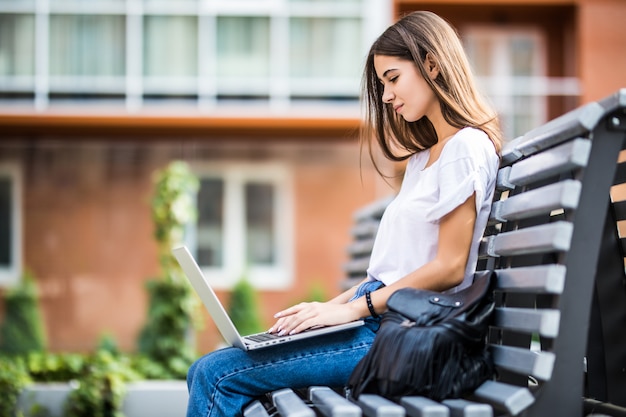 Heureuse femme tapant sur un ordinateur portable et regardant la caméra assise sur un banc à l'extérieur