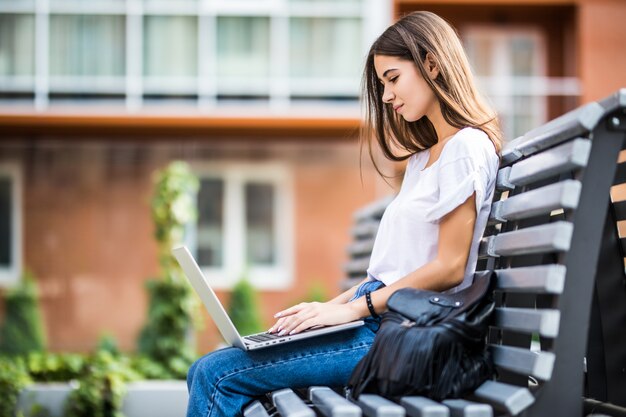 Heureuse femme tapant sur un ordinateur portable et regardant la caméra assise sur un banc à l'extérieur