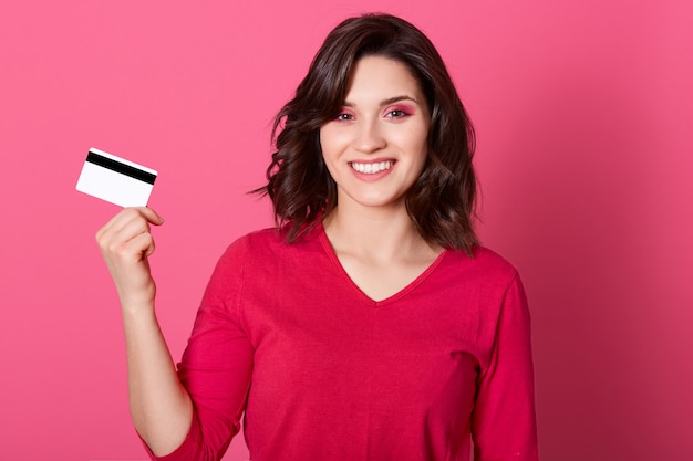 Heureuse femme tenant une carte de crédit dans les mains, regardant la caméra avec un sourire à pleines dents, ayant beaucoup d'argent pour les achats en ligne, portant une chemise décontractée rouge, a les cheveux noirs.