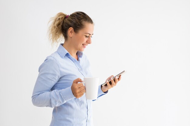 Heureuse femme tenant la tasse et lecture de message sur smartphone