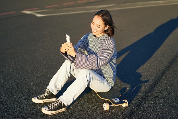 Heureuse fille asiatique assise sur une planche à roulettes prend selfie avec longboard fait des visages mignons journée ensoleillée à l'extérieur