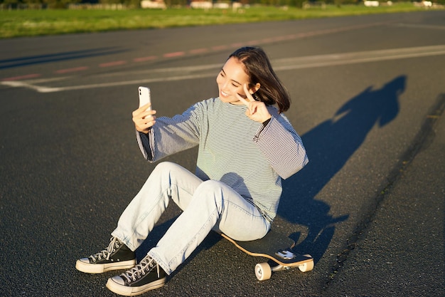 Heureuse fille asiatique assise sur une planche à roulettes prend selfie avec longboard fait des visages mignons journée ensoleillée à l'extérieur