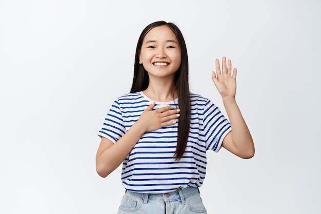 Heureuse fille asiatique souriante levant une main, posant la paume sur le cœur, jurant, faisant une promesse, se présentant, debout sur du blanc