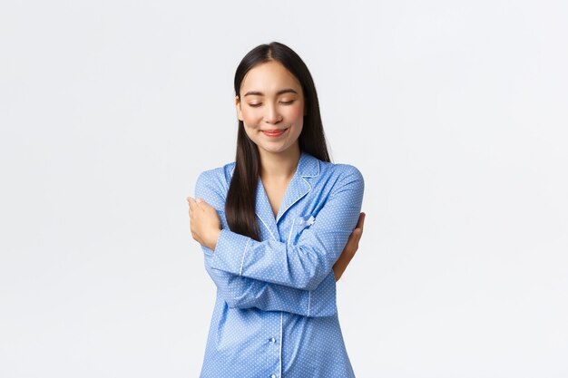 Heureuse fille asiatique tendre et rêveuse en pyjama bleu, fermer les yeux et sourire en rêvant, se serrant dans ses bras, embrassant son propre corps en pyjama, debout sur fond blanc détendu, rêvant