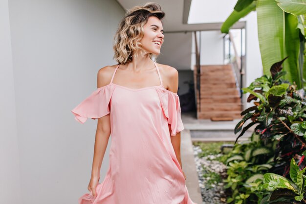 Heureuse fille aux cheveux courts porte des vêtements roses profitant d'une bonne journée. Photo extérieure d'un modèle féminin caucasien insouciant marchant à côté de plantes vertes dans la cour.