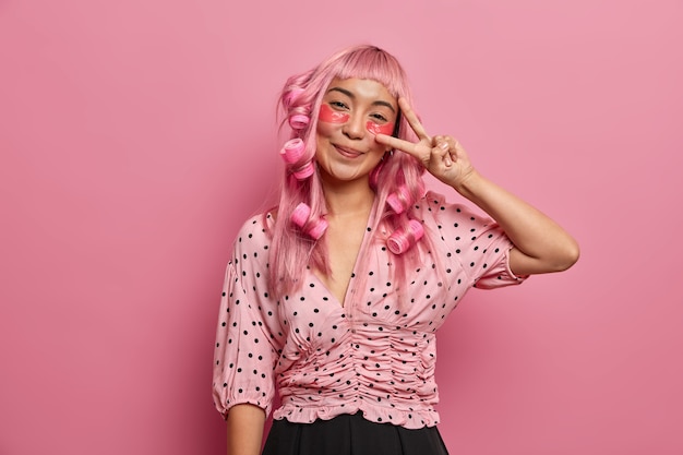 Heureuse fille aux cheveux roses applique des patchs oculaires pour réduire les cernes, fait un signe de paix, porte des bigoudis pour avoir des boucles parfaites